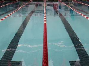 A close-up view of a competitive swimming pool from the pool deck's perspective, with a red and white lane rope in the center, converging neighboring lane lines, blue water, and black guidelines on the pool floor.